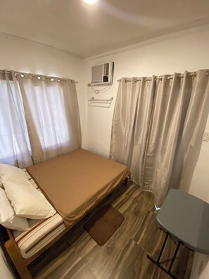 Une petite chambre avec un lit simple, un plancher en bois, une petite table et un climatiseur fixé au mur.