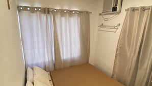 A small, single-bed room with a wooden headboard, a beige mattress, and a wooden floor. There is a window with curtains, a wall-mounted air conditioner, and a towel rack. - Simple, Practical & Comfy Room 4 (General Luna)