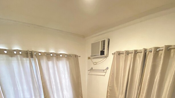 A small, single-bed room with a wooden headboard, beige curtains, a wall-mounted air conditioner, and a wooden floor. - Simple, Practical & Comfy Room 4 (General Luna)
