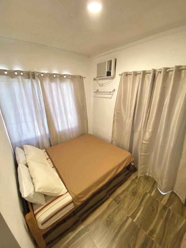 Une petite chambre avec un lit simple, une tête de lit en bois, des rideaux beiges, un climatiseur fixé au mur et un plancher en bois.