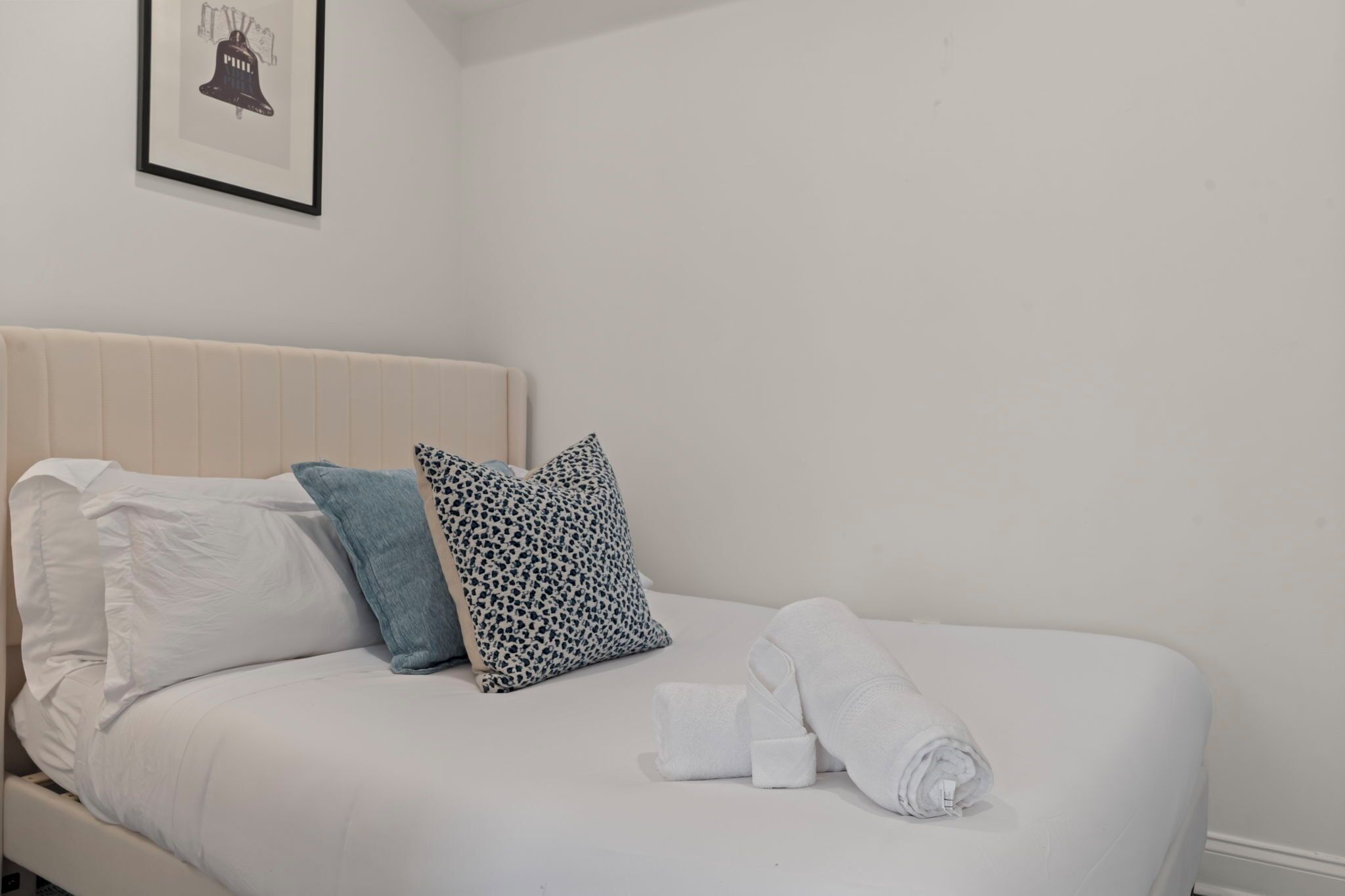 A neatly made bed with white and patterned pillows, a rolled white towel, and a framed picture on the wall.