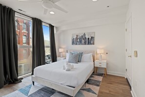 A modern hotel room with a large bed, a cityscape painting, a ceiling fan, and a view of a building through the window. - Apartmint at 116 Chestnut Philadelphia (Philadelphia)