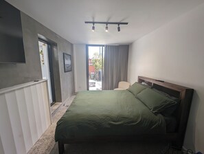 A bedroom with a bed, a TV mounted on the wall, a chair, and a view through a door to the outside.