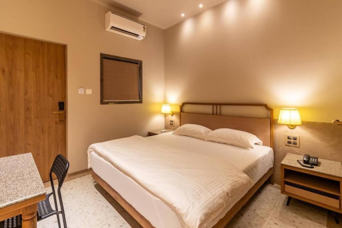 Una habitación de hotel con cama, lámparas de noche, escritorio, silla y aire acondicionado.