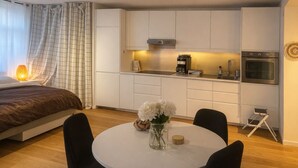 A modern bedroom with a bed, a bedside table with a lamp, and a view of a kitchenette with a coffee maker and oven.