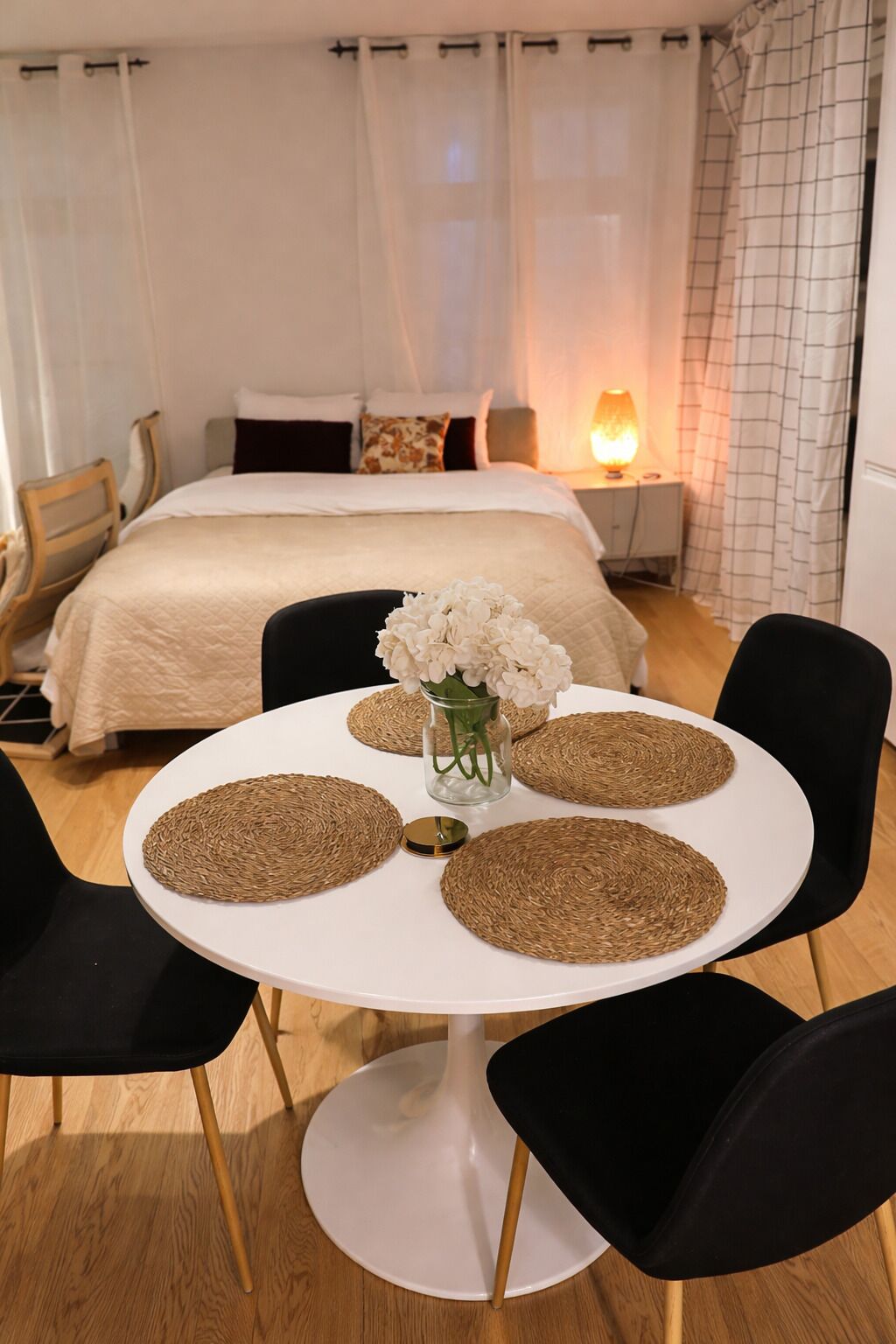 A hotel room with a bed, a bedside table with a lamp, a round table with woven placemats, and black chairs.
