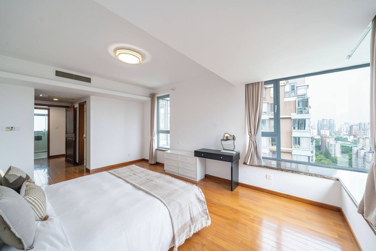 Un dormitorio con cama, escritorio, ventana con vistas a edificios y puerta que da a otra habitación.