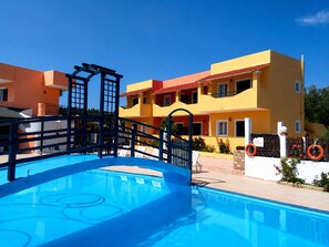 A swimming pool with a blue deck, a wooden fence, and a building with balconies and a red roof in the background. - Nikos Villas - Garden & Pool View Gem in Kavos (Corfu)