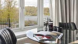 A room with a glass table, a book, a coffee maker, and a view of trees through large windows. - Lakeside Studio Ridgestone (Windermere)