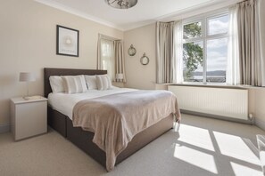 Um quarto com cama, mesas de cabeceira, janela com cortinas, radiador e uma imagem emoldurada na parede.