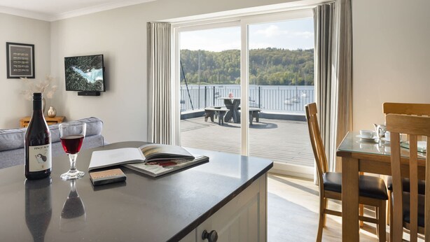 A room with a dining table, chairs, a glass-top table, a bottle of wine, a glass of red wine, a book, and a view of a lake through large windows. - Mereside Apartment Ridgestone (Windermere)