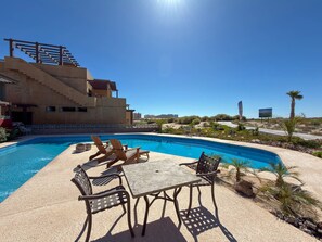 Una piscina con sillones de descanso y una mesa, rodeada por un paisaje desértico y un edificio con escaleras.