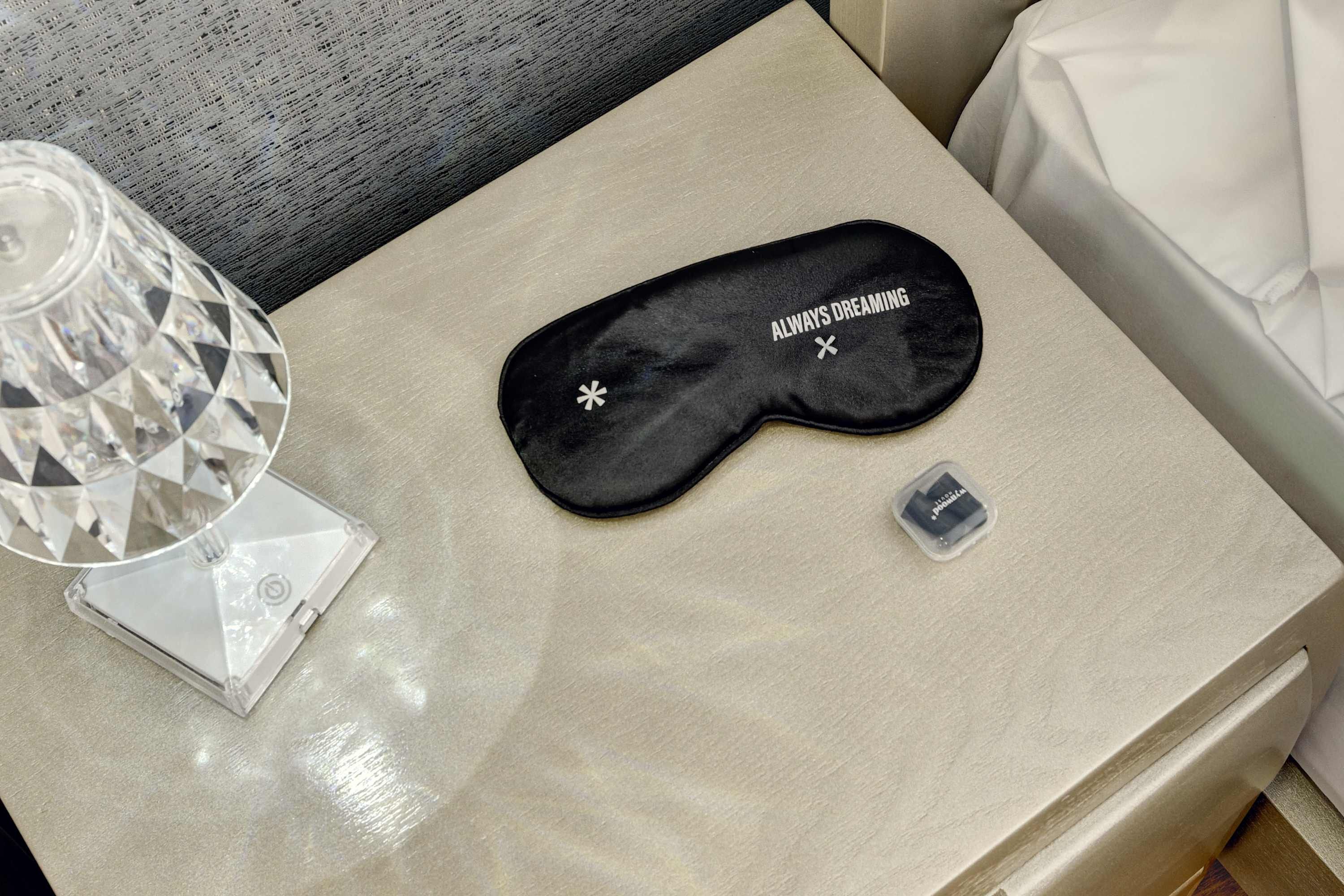 Une table de chevet avec un masque de sommeil noir, un petit récipient en plastique et une lampe en verre transparent.