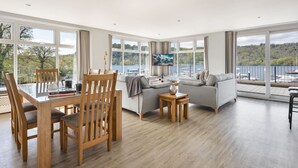 Ein großzügiger Wohnraum mit Essbereich, einem grauen Sofa und einem Fernseher. Große Fenster bieten Blick auf den See und Boote.