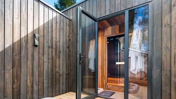 Uma área de spa em madeira com hidromassagem, portas de vidro que dão para um vestiário e um deck de madeira.