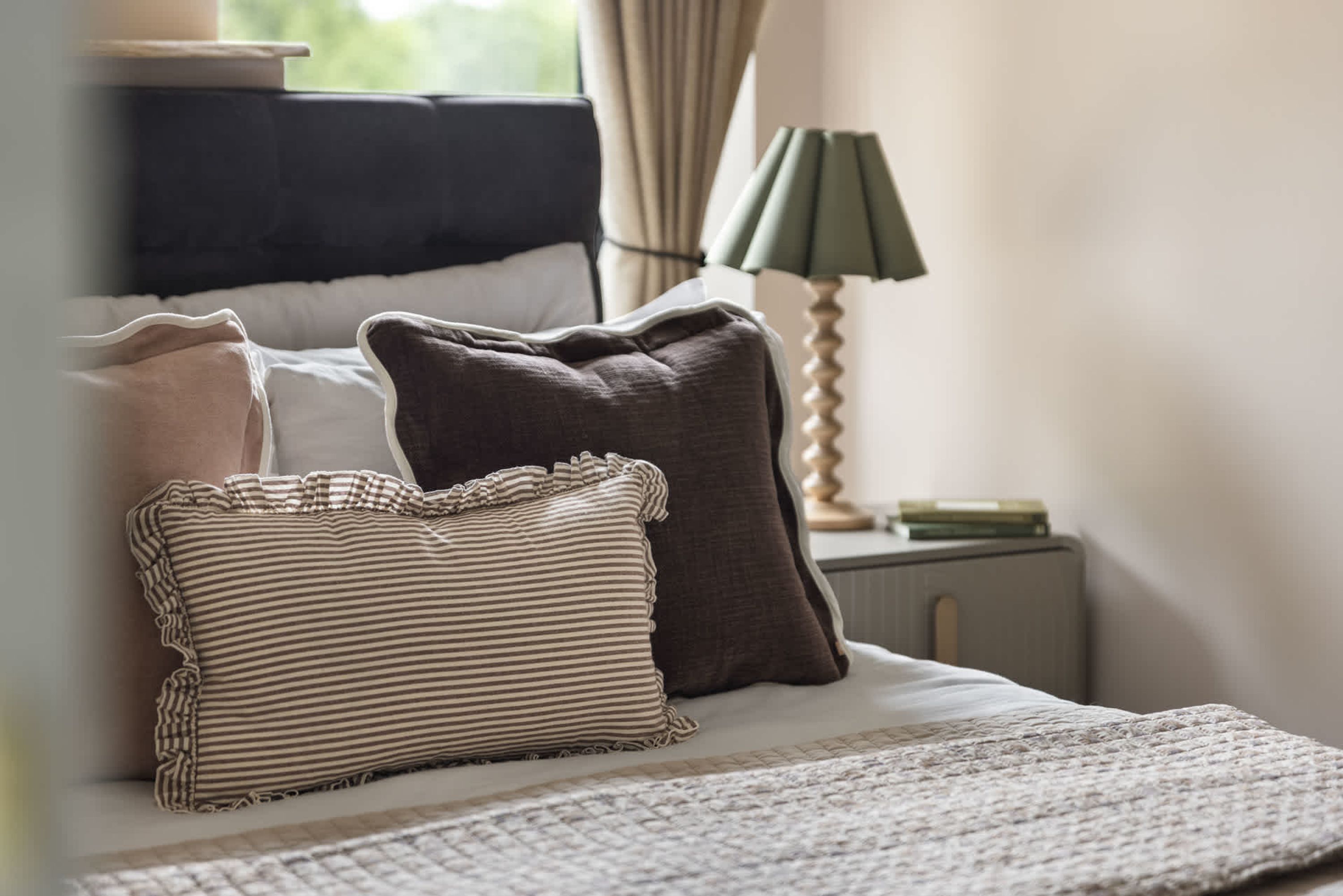 A neatly made bed with striped and solid pillows, a bedside lamp, and a small bedside table with a book.