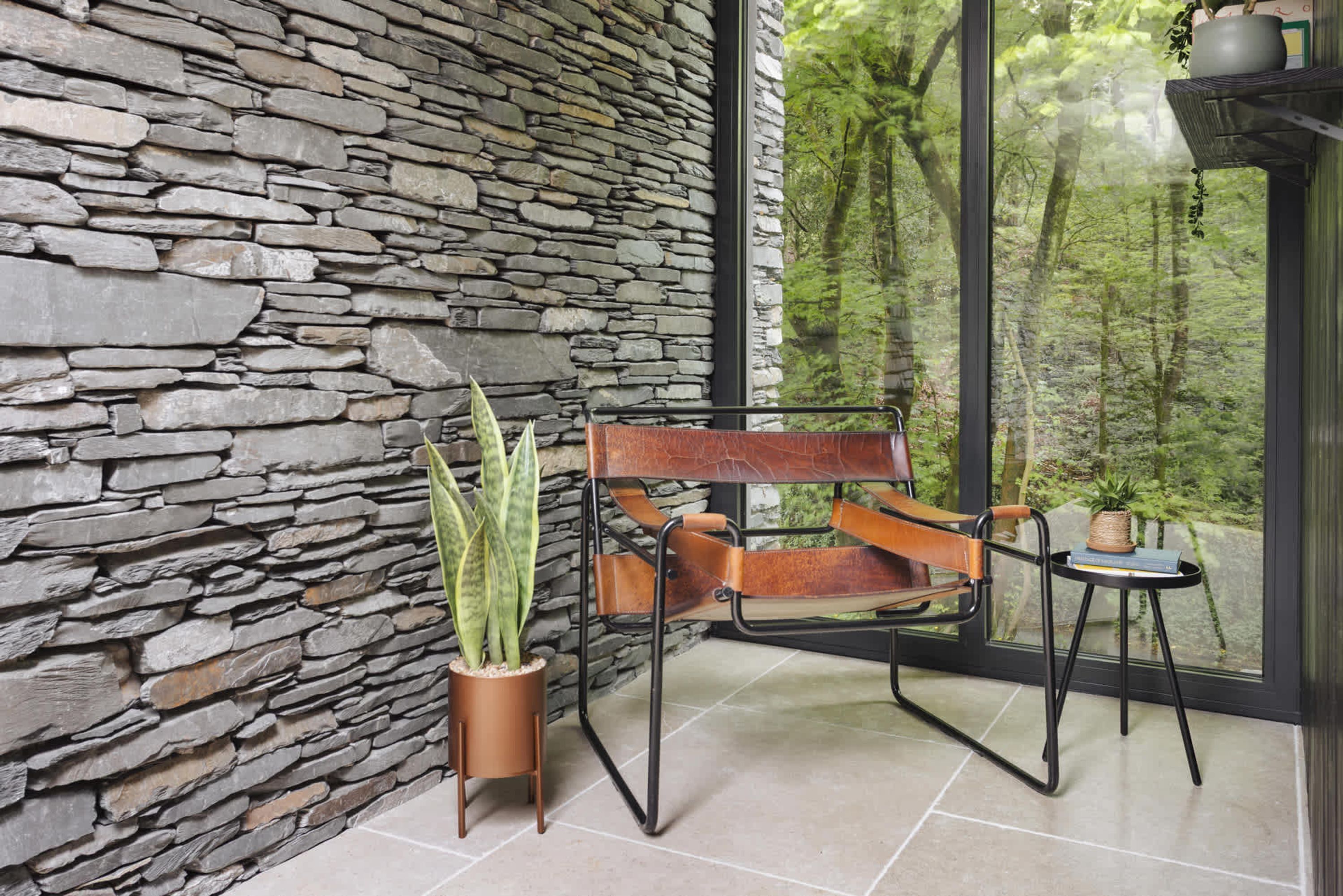 Una habitación con pared de piedra, un ventanal con vistas a un bosque, un sillón de cuero y una mesita con una planta.