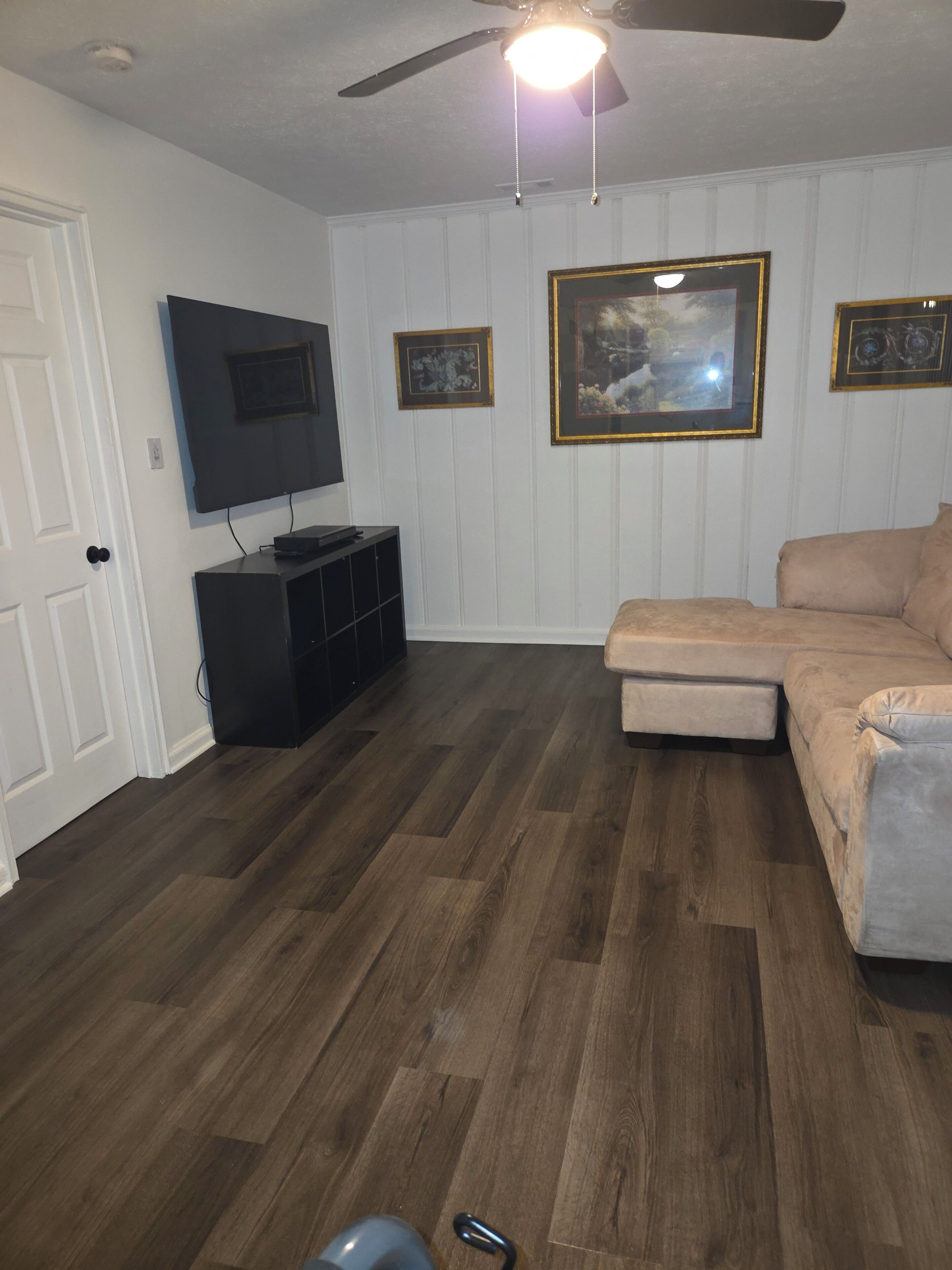 A living room with a ceiling fan, a flat-screen TV, a beige sofa, and a wooden floor.