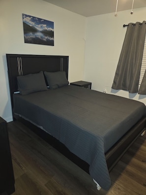 Un dormitorio con cama, una mesita de noche, un cuadro colgado en la pared y una ventana con cortinas.