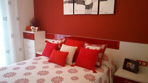 Quarto com cama, almofadas, mesa de cabeceira, cômoda e uma parede com obras de arte.