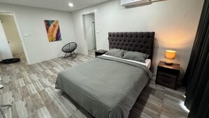 A modern bedroom with a large bed, a chair, a bedside table with a lamp, and an air conditioning unit.