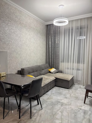 Una sala de estar moderna con un sofá seccional, una mesa de comedor con sillas y un amplio ventanal con cortinas ligeras.