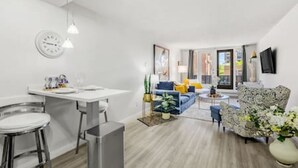 A modern living room with a sofa, a dining area with a table and chairs, and a kitchen area with a clock and a plant. - Bright Beltline Suite, Sleeps 4, Free Parking (Calgary)