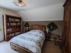 A bedroom with a wooden bed, a bookshelf, a wall-mounted lamp, and a decorative wreath. - Gite La thébaïde (Thoury)