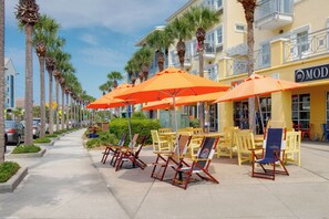 A sunny street with outdoor seating, palm trees, and a yellow building with a sign. - Bluefish House • Six-Seater Golf Cart • Walk to Beach • Private Hot Tub (Santa Rosa Beach)