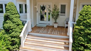 Ett vitt hus med en veranda av trä, en krukväxt och en stol.