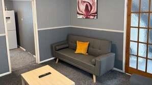 A room with a grey sofa, a small wooden table, a remote control, a refrigerator, and a window with a view. - Blyth City Delight Free Parking (Blyth)