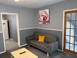 Ein Zimmer mit grauem Sofa, einem kleinen Holztisch, einer Fernbedienung, einem Kühlschrank und einem Fenster mit Blick.