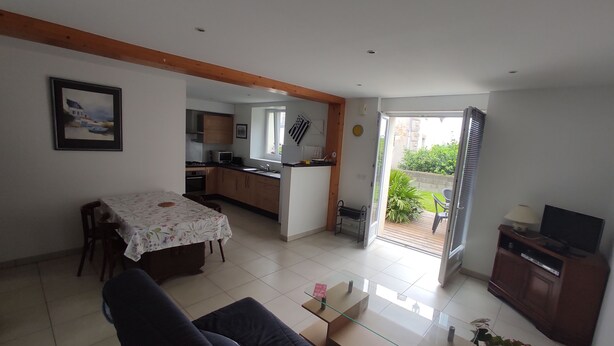 Een woonkamer met een bank, een glazen salontafel, een eettafel met een bloemen tafelkleed, een keuken met houten kasten en een schuifdeur die leidt naar een terras met tuinmeubilair.