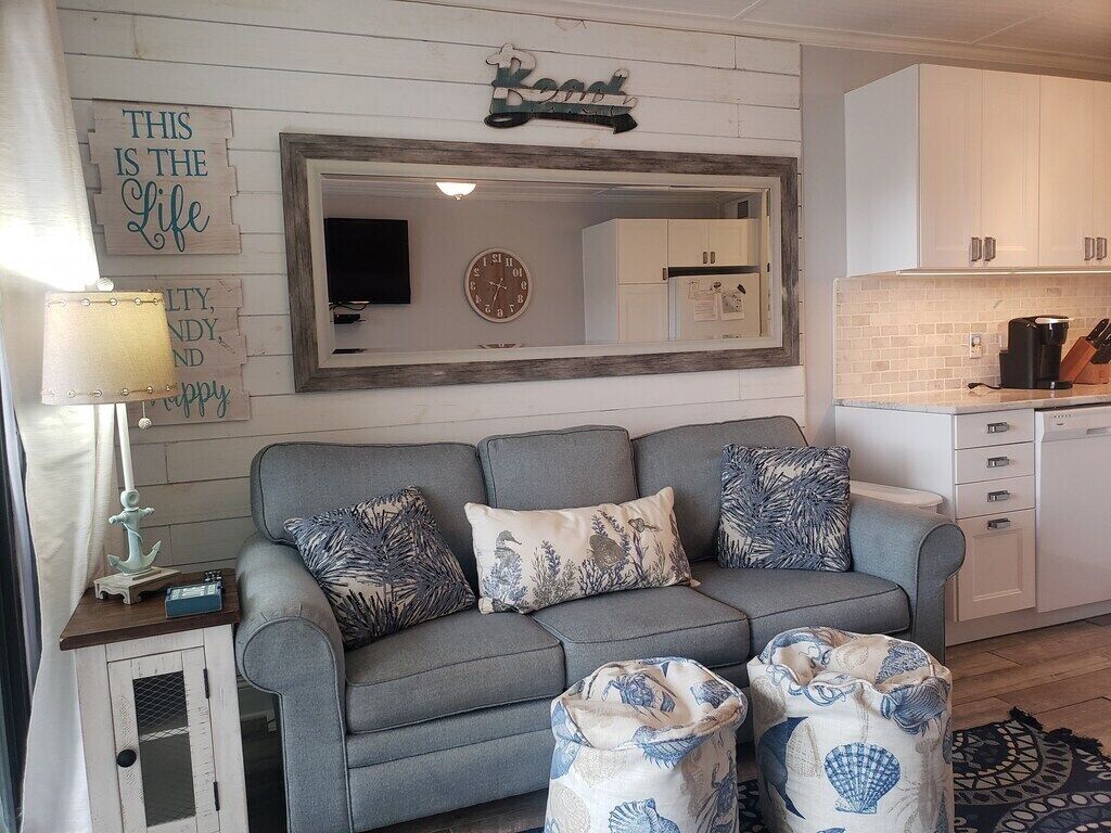 A living room with a grey sofa, decorative pillows, a mirror, and a kitchen area with white cabinets and a coffee maker.