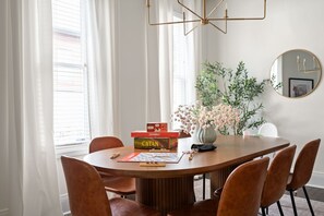 Uma sala de jantar com uma mesa redonda, cadeiras, um lustre, um vaso com flores e um espelho na parede.
