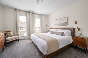A bedroom with a large bed, a bedside table, a nightstand, a chair, and a window with curtains. - Walk to Mass Ave: 2 homes, hot tub, private yard (Indianapolis)