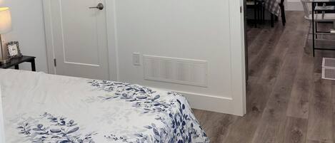 Een slaapkamer met een bed, een deur, een ventilatie rooster en een gang die leidt naar een eetruimte.