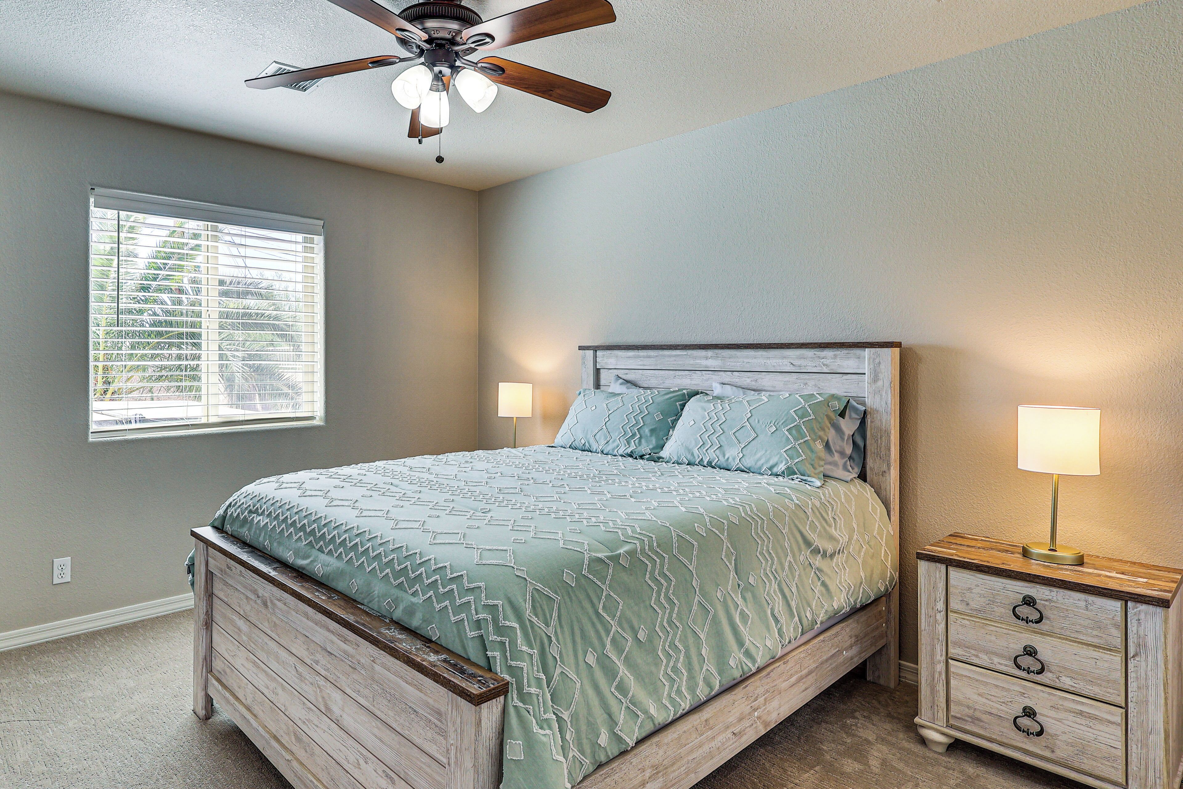 A bedroom with a bed, a nightstand, a lamp, a ceiling fan, and a window with blinds.