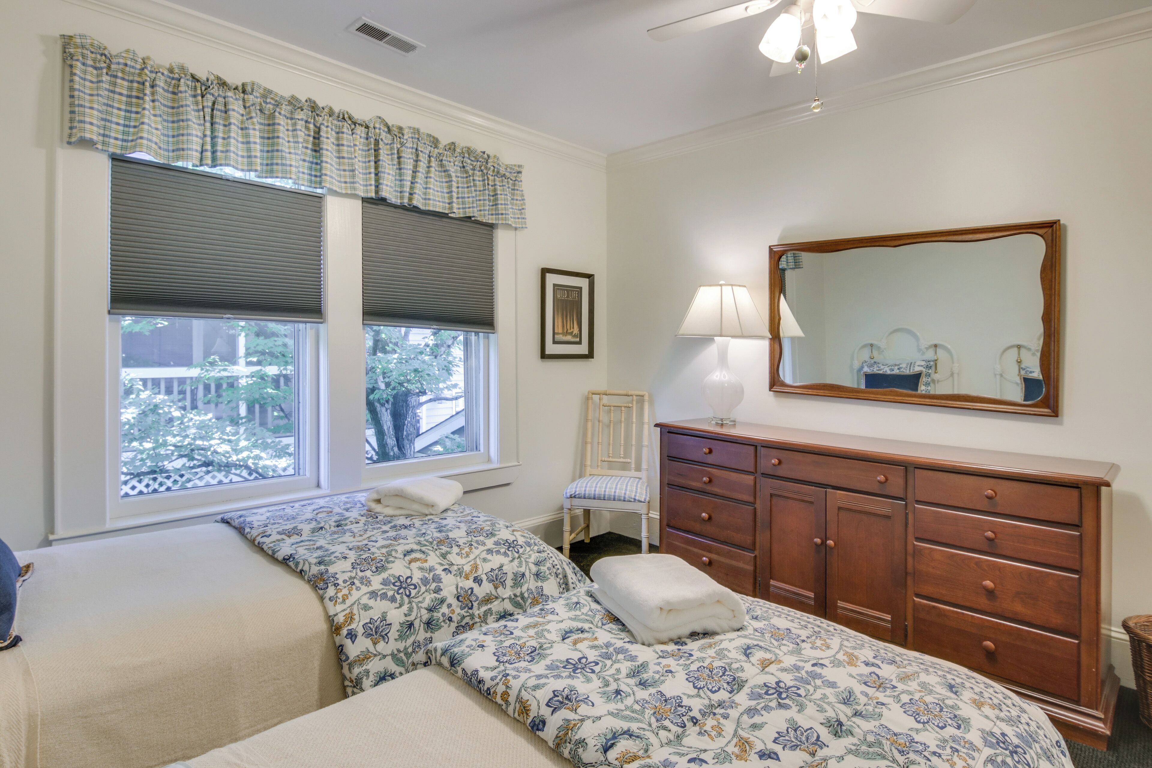 A bedroom with two beds, a dresser, a chair, a window with blinds, a lamp, and a mirror.