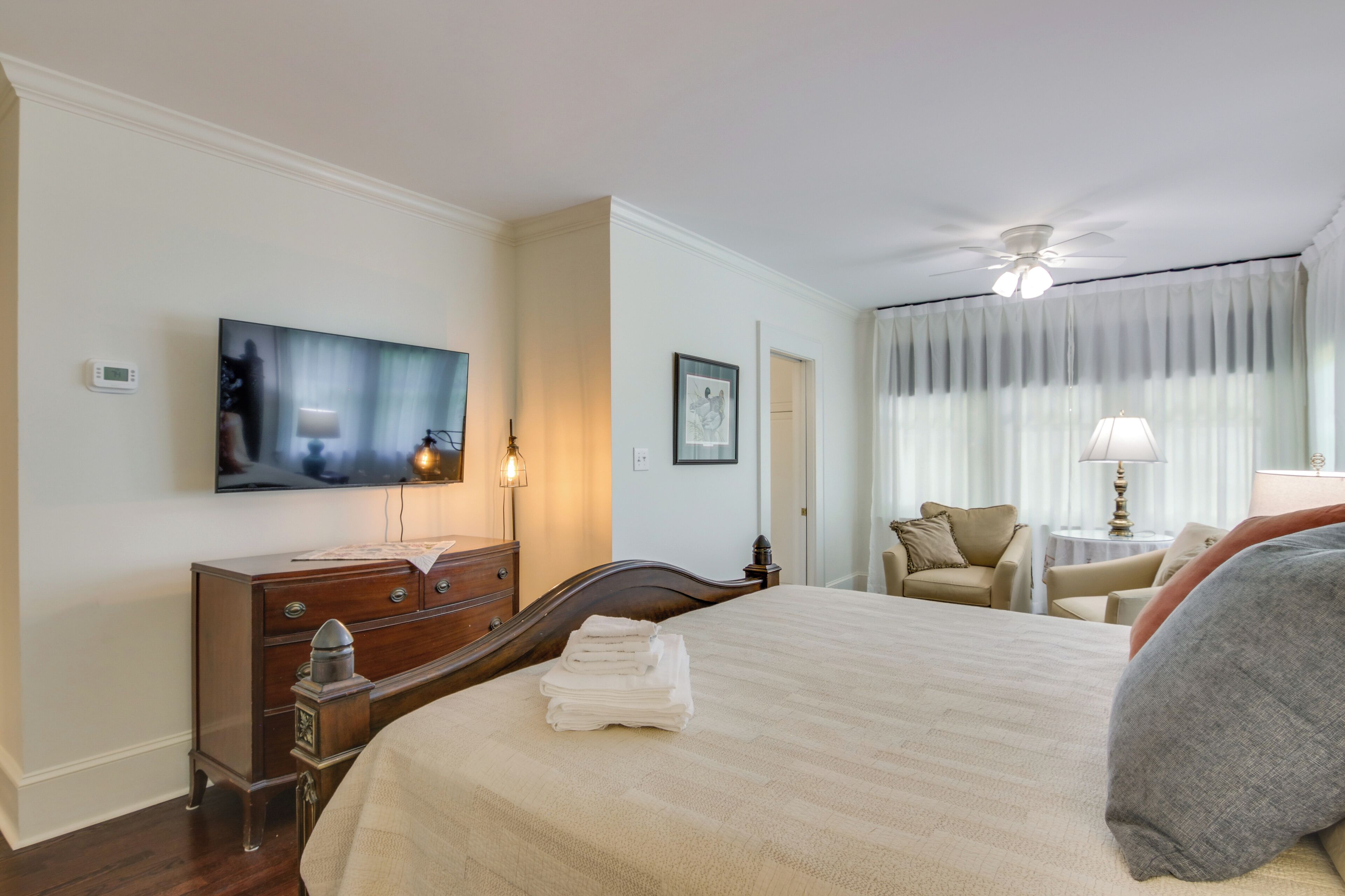 Ein Hotelzimmer mit Bett, Kommode, Fernseher, Deckenventilator und einem fensterbepflanzten Fenster.