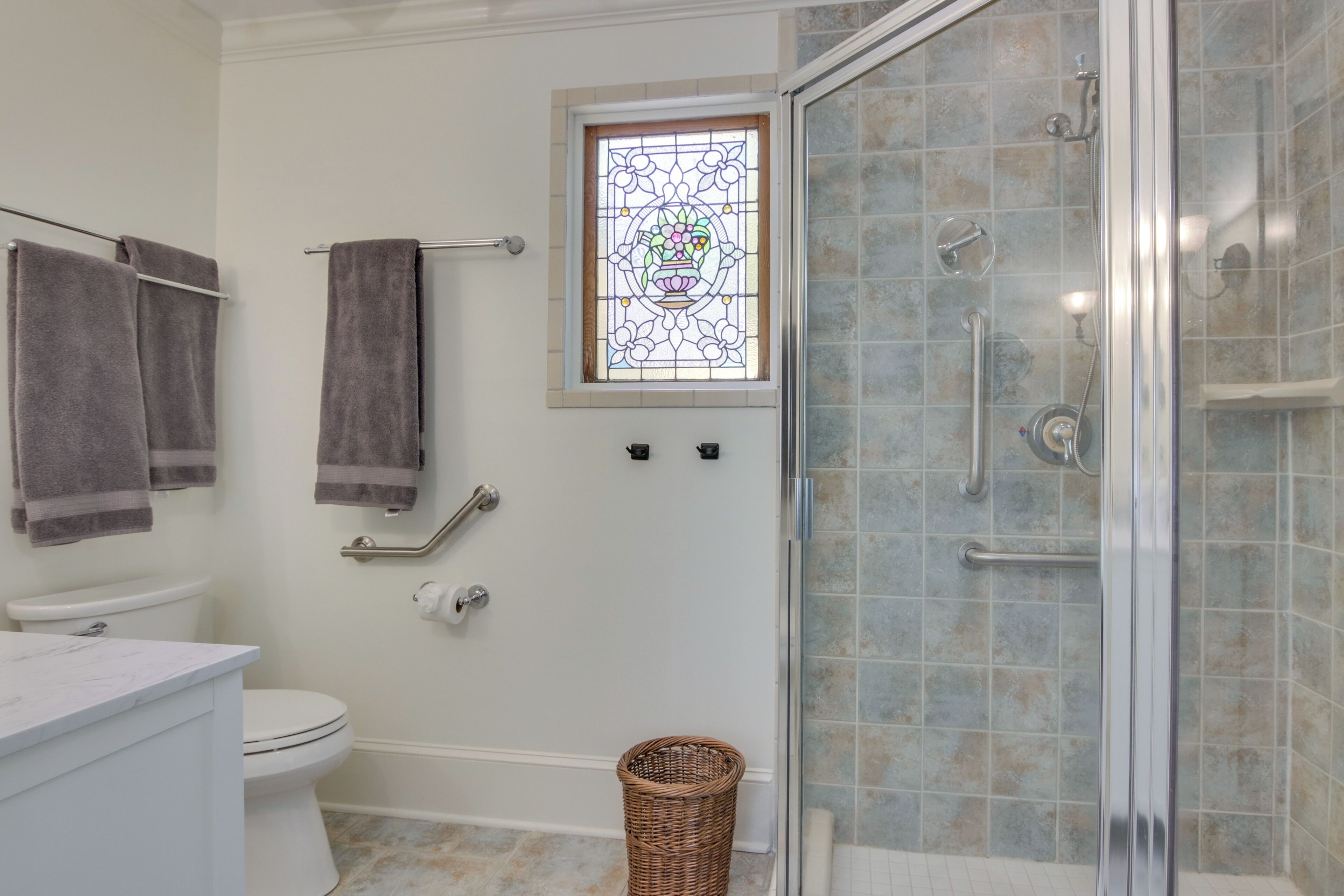 A bathroom with a toilet, a shower with glass doors, a white sink, and a window with a stained glass design.