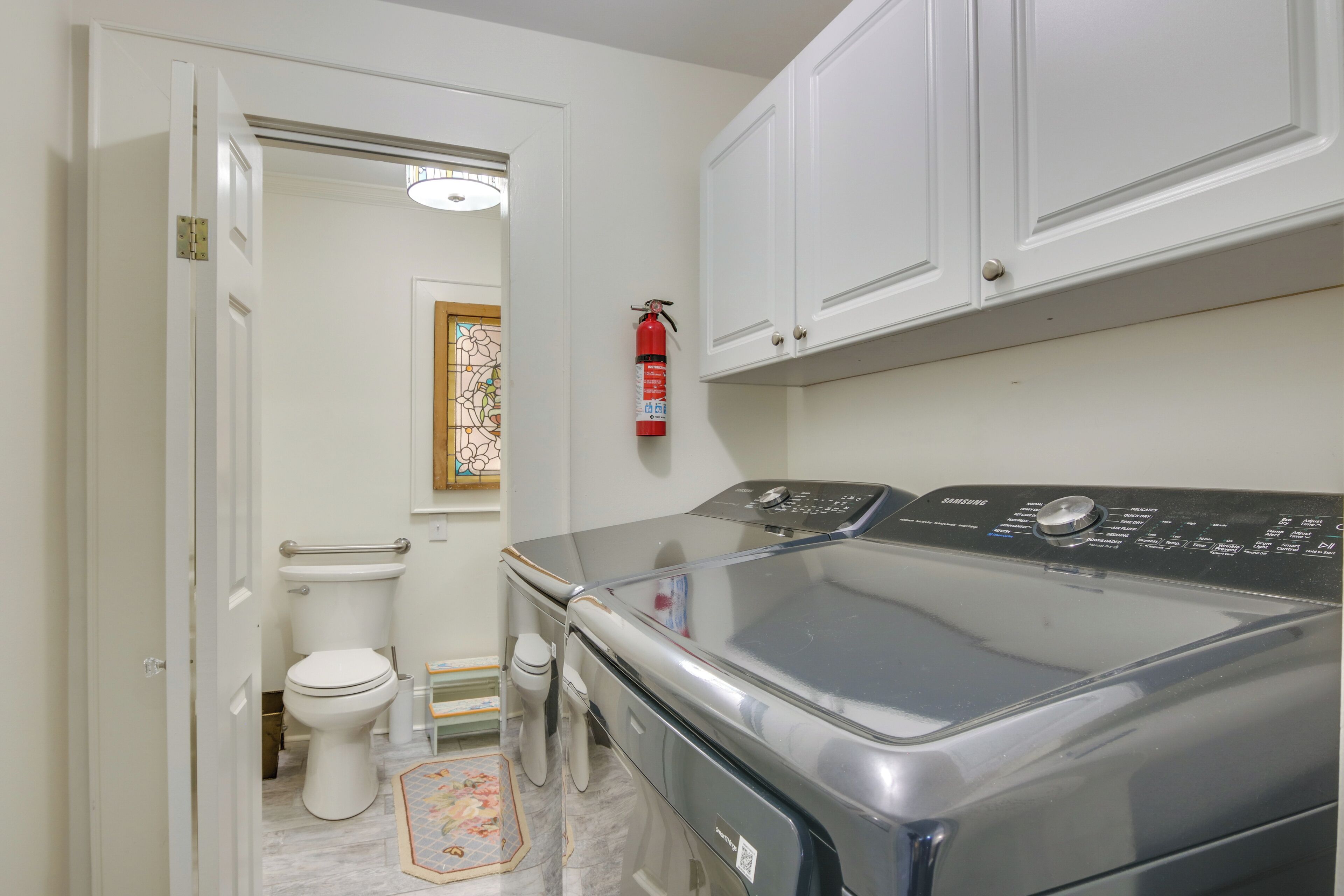 A laundry room with a front-loading washer and dryer, a white toilet, a fire extinguisher, and a small rug on the floor.