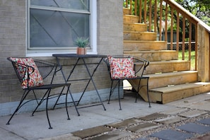 A patio with two metal chairs and a table, a wooden staircase, and a brick wall. - Home w/ Fenced Yard, 13 Mi to Kansas City Stadium! (Kansas City)