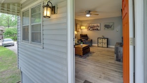 A covered porch with a sliding door leading to a living room with a ceiling fan, a sofa, a coffee table, and a rug. - KC Stadium Gateway: Home w/ Private Yard (Kansas City)