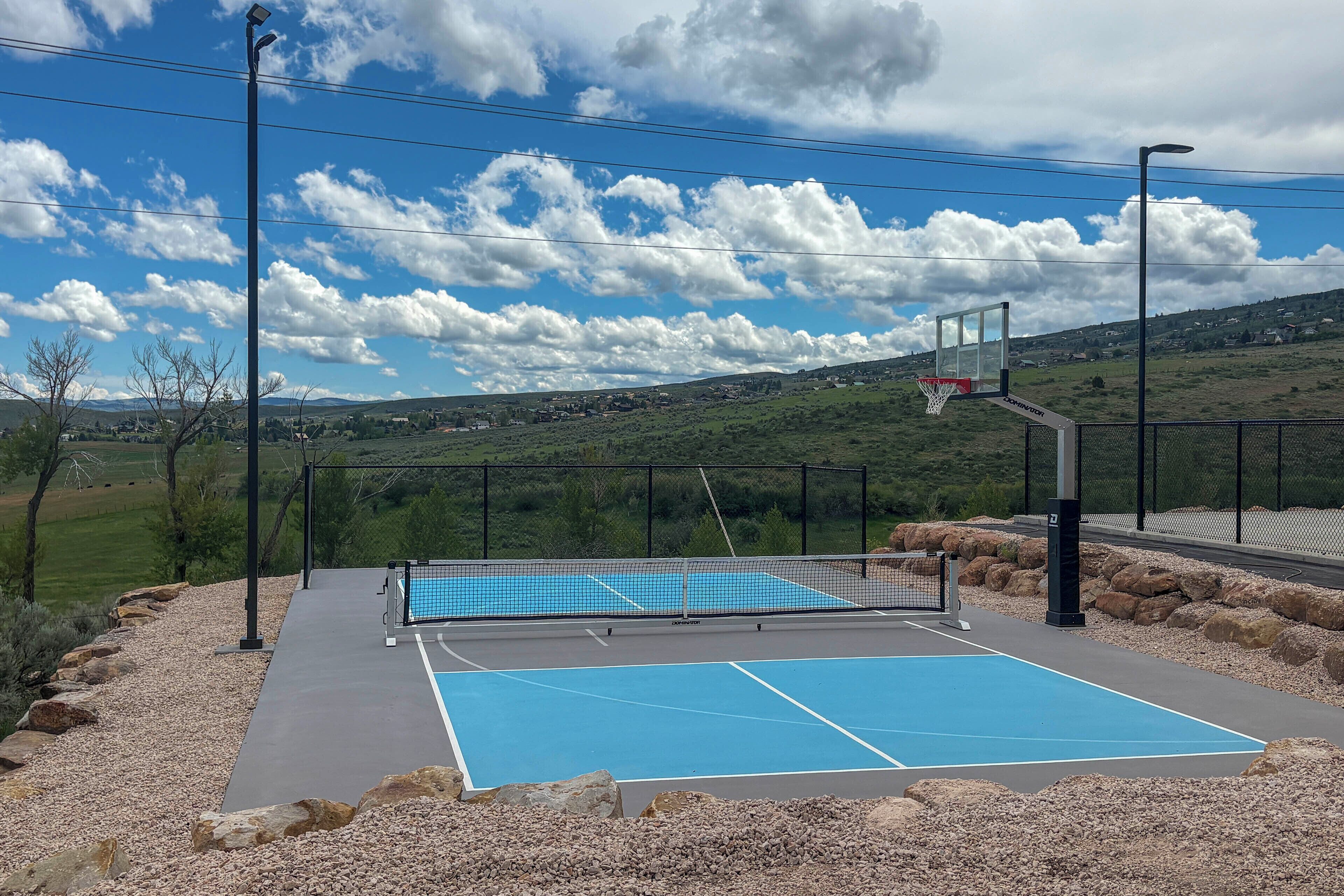 Una cancha de baloncesto al aire libre con aro, rodeada por una valla, y una vista despejada de un paisaje montañoso al fondo.