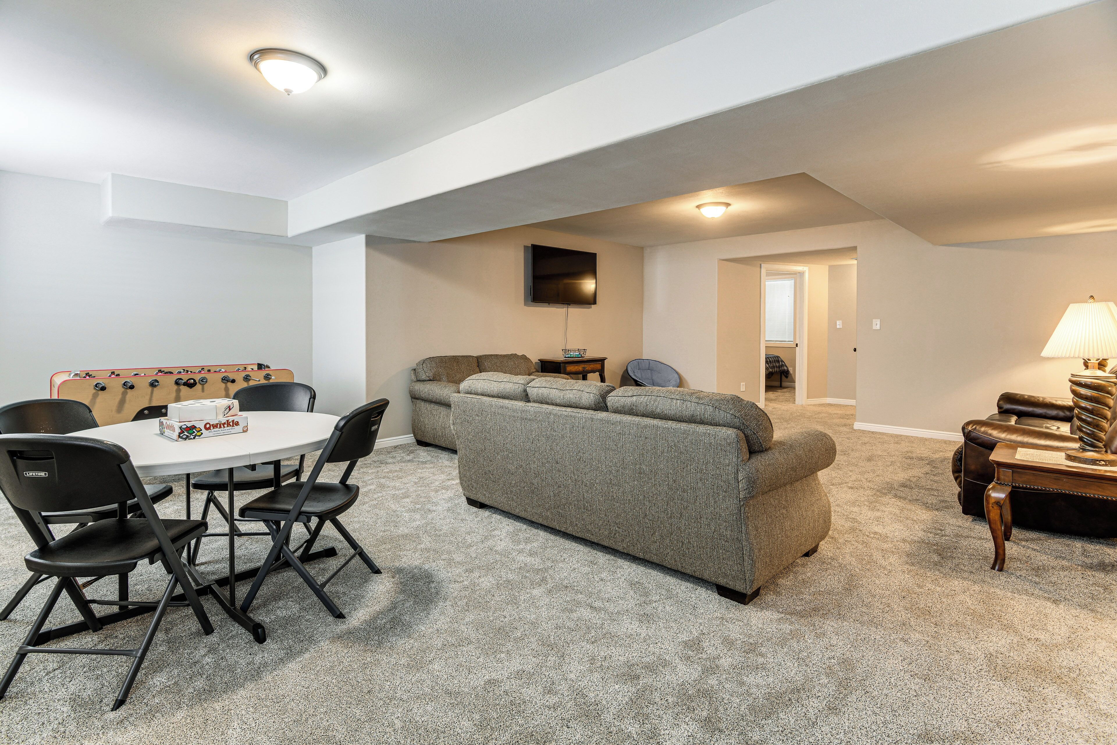 A living room with a gray sofa, a round table with chairs, a flat-screen TV mounted on the wall, and a lamp on a side table.