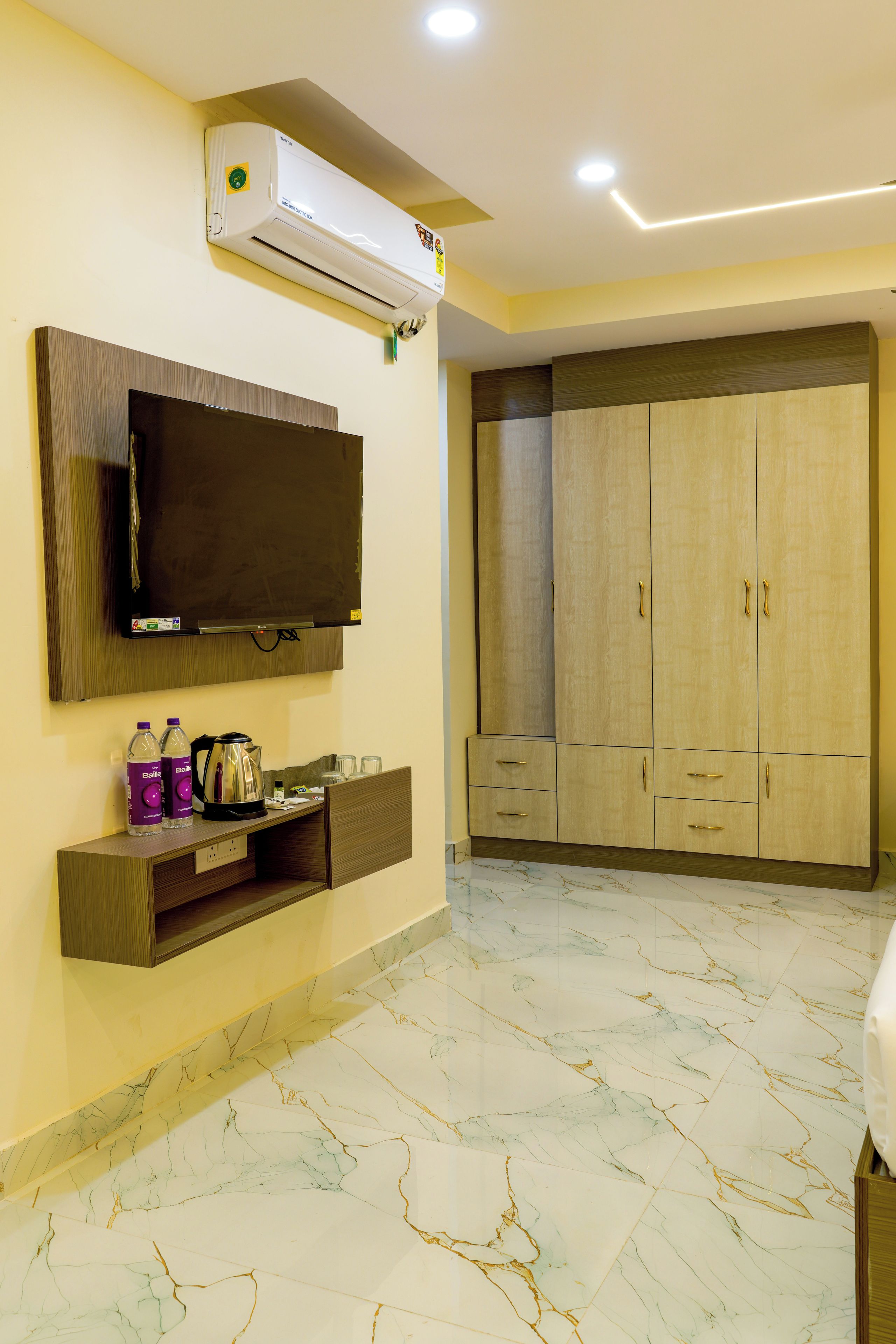 A hotel room with a flat-screen TV mounted on the wall, a minibar, a wardrobe with multiple doors, and a marble floor.