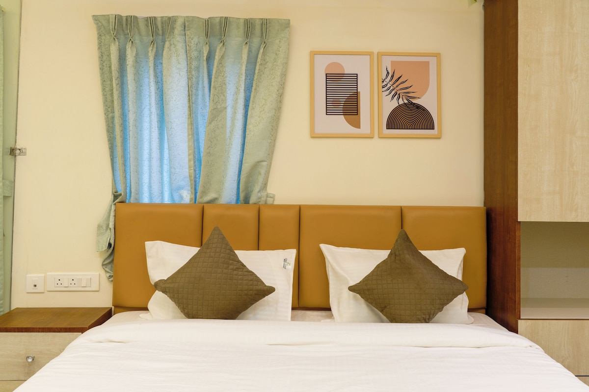 A hotel room with a bed, two pillows, a headboard, a nightstand, a wall socket, and two framed artworks.