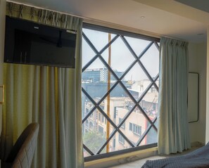 A room with a large window featuring a diamond-patterned grille, curtains, a bed, and a television on the wall.