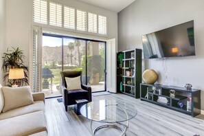 Ein Wohnzimmer mit einer Couch, einem Glastisch, einem Sessel, einem Bücherregal und einem an der Wand montierten Fernseher.